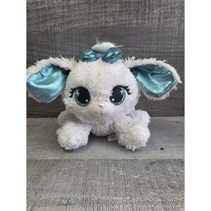 May include: A white plush puppy with large, sparkling blue eyes and matching ears and bow. The stuffed animal has a fluffy texture and is adorned with silver glitter accents. The puppy is sitting on a gray wooden surface.