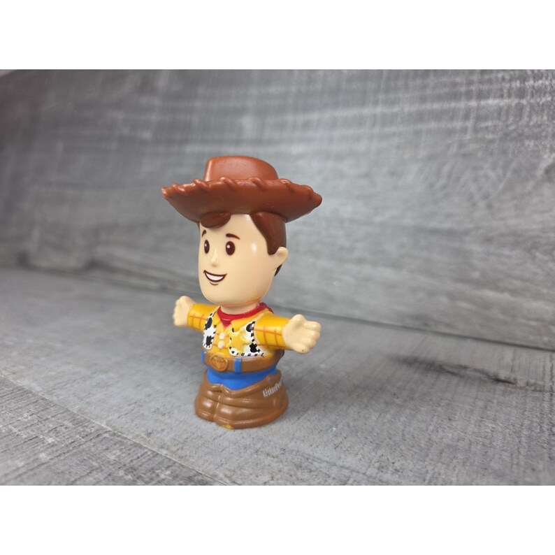 May include: A small plastic toy figure of a cowboy character, wearing a brown hat, yellow shirt, blue pants, and brown boots. The figure has outstretched arms and a smiling face. The toy is a collectible item.