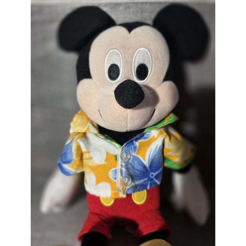 Puede incluir: Un peluche de Mickey Mouse con una camisa hawaiana colorida con estampados florales amarillos, azules y blancos. El mu&ntilde;eco tiene orejas negras, una nariz negra y pantalones cortos rojos. La camisa del mu&ntilde;eco est&aacute; desabrochada, revelando un cuello verde.