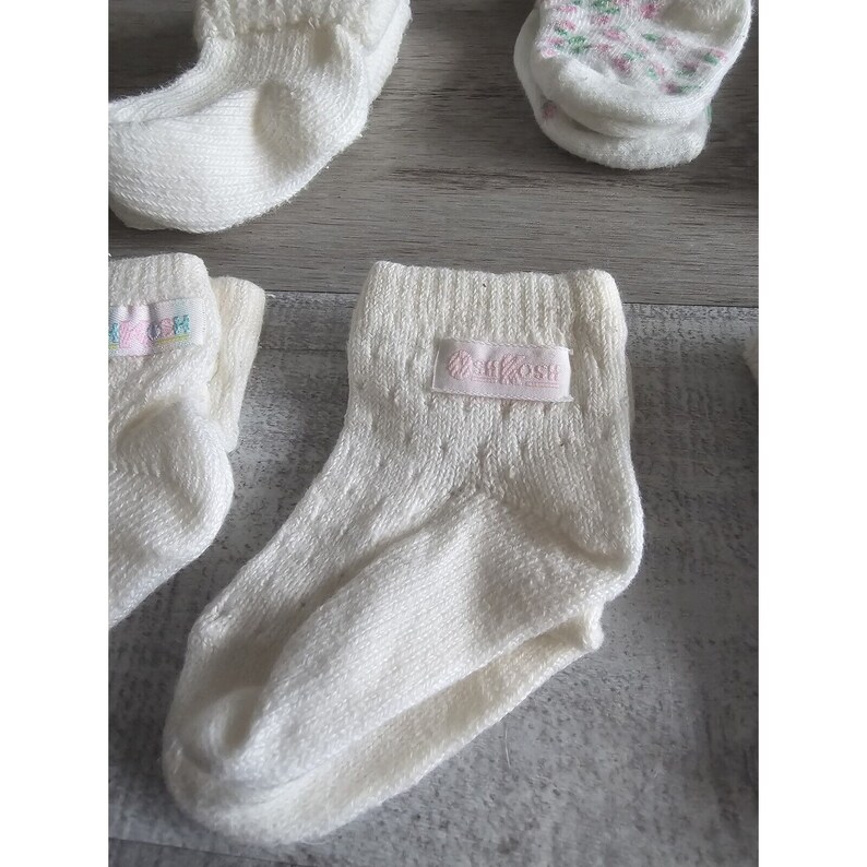 Puede incluir: Un conjunto de calcetines blancos para beb&eacute; con una etiqueta rosa que dice " OshKosh" en un calcet&iacute;n.