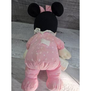 Puede incluir: Mu&ntilde;eca de peluche de Minnie Mouse, vista desde atr&aacute;s, vestida con un mono rosa con un estampado floral blanco. La mu&ntilde;eca tiene orejas negras, un lazo rosa y un cuello de encaje blanco. La mu&ntilde;eca sostiene un peque&ntilde;o objeto blanco.