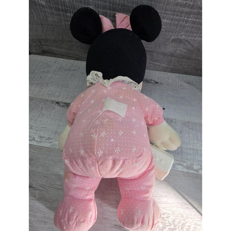Puede incluir: Mu&ntilde;eca de peluche de Minnie Mouse, vista desde atr&aacute;s, vestida con un mono rosa con un estampado floral blanco. La mu&ntilde;eca tiene orejas negras, un lazo rosa y un cuello de encaje blanco. La mu&ntilde;eca sostiene un peque&ntilde;o objeto blanco.