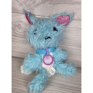 May include: A plush toy dog with shaggy light blue fur, large dark eyes, and pointed ears featuring sparkly pink inner linings. The toy wears a light blue plastic collar with a round purple tag that has a white heart icon and a blank name space.