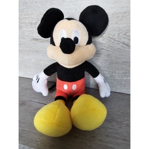 Puede incluir: Un juguete de peluche de Mickey Mouse con orejas negras, cuerpo negro, pantalones cortos rojos con botones blancos y zapatos amarillos.