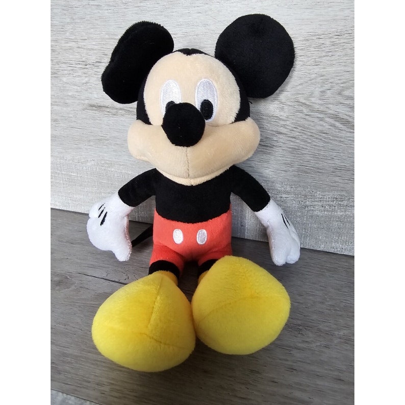 Puede incluir: Un juguete de peluche de Mickey Mouse con orejas negras, cuerpo negro, pantalones cortos rojos con botones blancos y zapatos amarillos.