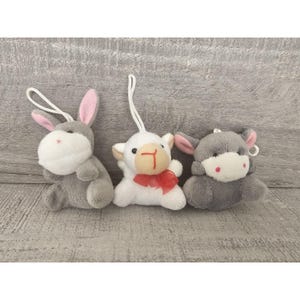 May include: Three plush toys: a gray rabbit, a white lamb with a red bow, and a gray cow. The toys are all small and have a soft, fuzzy texture.