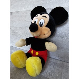 May include: A plush black and yellow Mickey Mouse toy with red shorts and black shoes. The toy is sitting on a light grey surface.