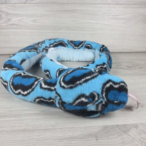 May include: A plush snake toy featuring a vibrant blue, black, and white patterned fur exterior. The stuffed animal is coiled in a circular shape and includes a small, light pink fabric tongue protruding from the front of its rounded head.