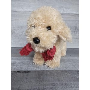 Puede incluir: Un perro de peluche con una bufanda roja. El perro es de color crema y tiene una textura esponjosa.