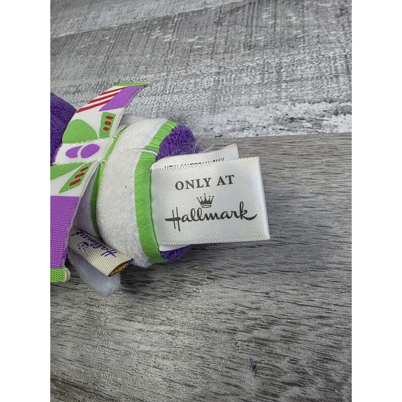 May include: A plush toy featuring a purple and white design with green accents. The toy has a tag that reads "ONLY AT Hallmark". The toy has a colorful design with a striped pattern.