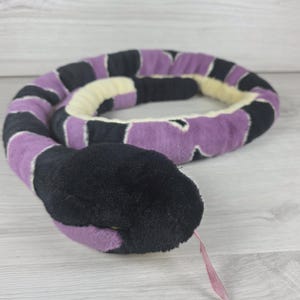 Puede incluir: Un juguete de serpiente de peluche con un patrón repetitivo de segmentos negros y morados a lo largo del cuerpo. La cabeza es de color negro sólido con una lengua de cinta rosa fina y pequeña que sobresale de la boca. El vientre es crema.