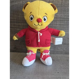 Puede incluir: Un peluche de tigre con pelaje amarillo, orejas marrones y una sudadera roja con capucha. El tigre lleva zapatillas rosas y blancas. El juguete tiene una expresión amigable con ojos grandes y una nariz pequeña.