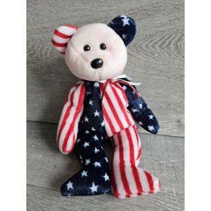 May include: A plush teddy bear with a red, white, and blue American flag pattern. The bear has a pink nose and black eyes.