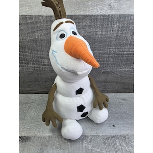 May include: A plush Olaf doll from the movie Frozen. The snowman has a white body, an orange carrot nose, and black eyes. His arms and hair are brown. He has a smiling expression and is sitting down.