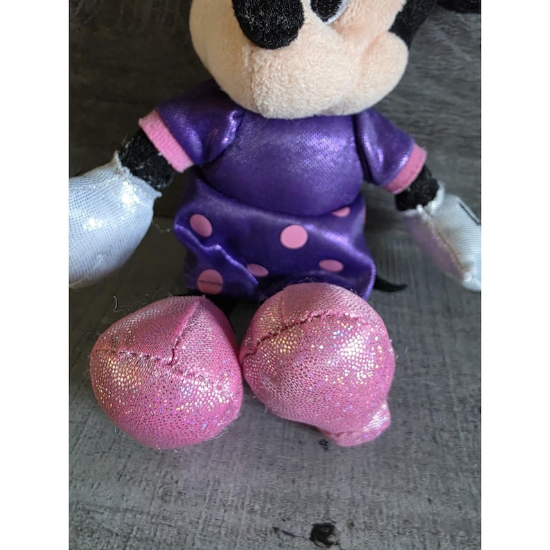 Op de afbeelding: Een pluche Minnie Mouse-pop met een paarse jurk met roze stippen. De pop heeft roze glitterschoenen, witte handschoenen en een zwarte kop met grote oren. De armen en benen zijn aan het lichaam bevestigd.