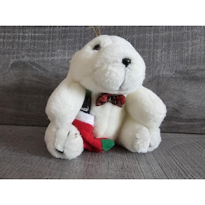 May include: A white plush polar bear Christmas ornament with a red and green plaid bow tie and a red and white stocking. The bear is holding a miniature Coca-Cola bottle and a red and white stocking.