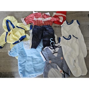 Pode incluir: Uma coleção de roupas de boneca vintage em vários estilos e cores. Inclui um casaco com capuz amarelo, camisa azul, calças de ganga, um conjunto de estilo marinheiro e macacões brancos. Os conjuntos apresentam detalhes como acabamentos em renda e fechos de botões.