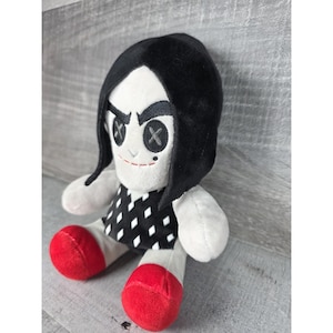 May include: A soft plush doll with a white face, black hair, and a black and white patterned dress. The doll has button eyes with an X design and red shoes. The doll is a soft toy.
