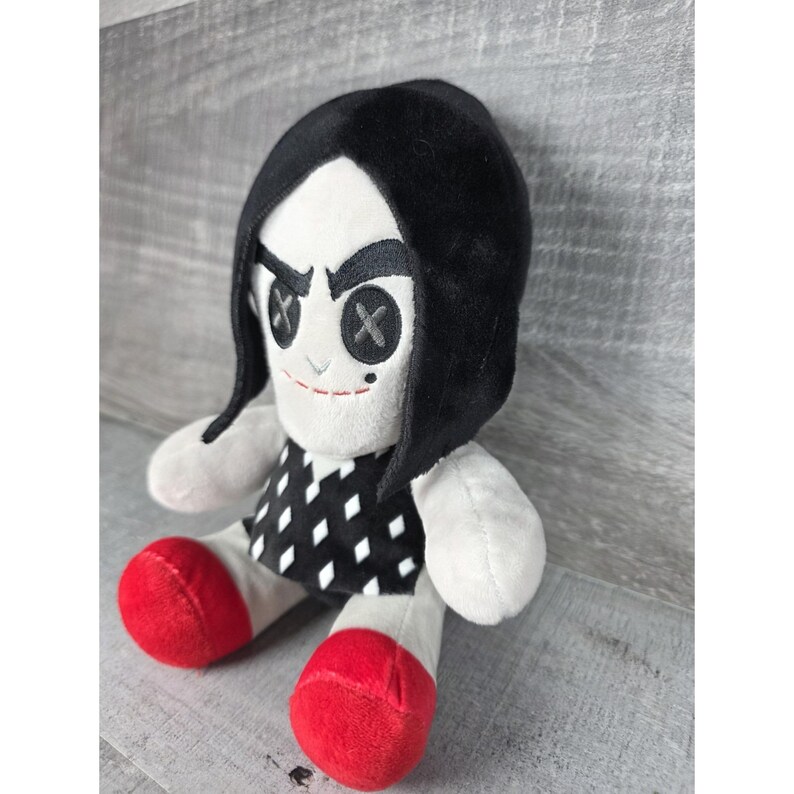 May include: A soft plush doll with a white face, black hair, and a black and white patterned dress. The doll has button eyes with an X design and red shoes. The doll is a soft toy.