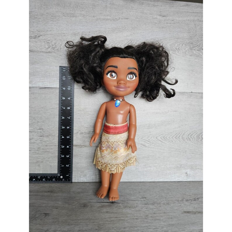 May include: A brown-skinned doll with black curly hair, wearing a brown and gold skirt with a red sash and a blue necklace. The doll has brown eyes and is wearing a light brown skirt with a red sash.