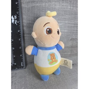May include: A plush baby doll with a light skin tone. The doll is wearing a white shirt with a bear graphic, blue sleeves, and a yellow bottom. It has a small yellow tuft of hair and brown eyes. A tag is attached.
