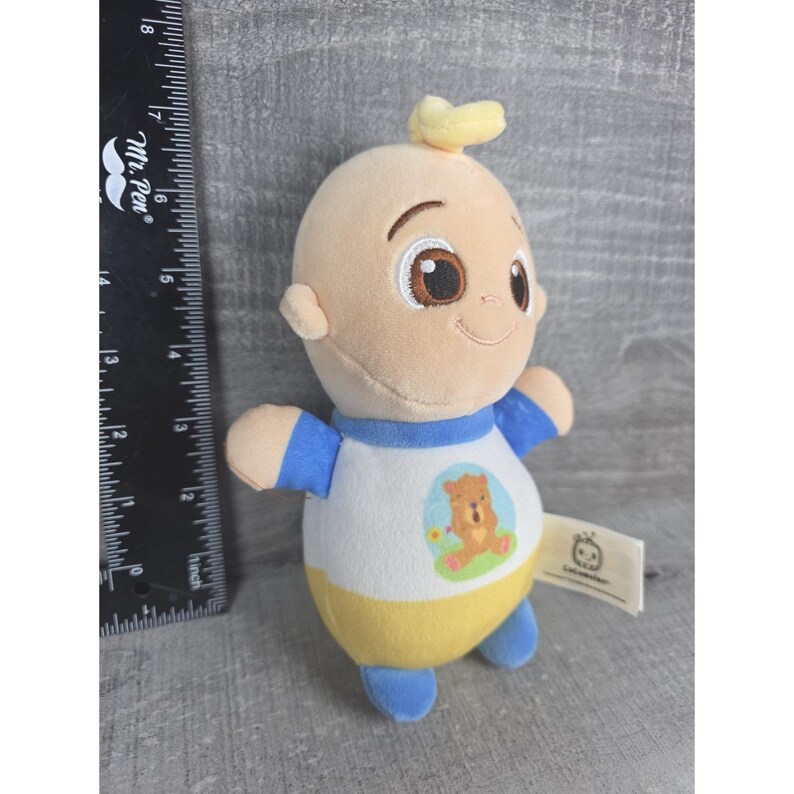 May include: A plush baby doll with a light skin tone. The doll is wearing a white shirt with a bear graphic, blue sleeves, and a yellow bottom. It has a small yellow tuft of hair and brown eyes. A tag is attached.