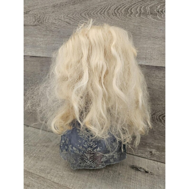 May include: A ceramic gnome figurine with a long, blonde wig. The gnome is wearing a blue and green patterned outfit.