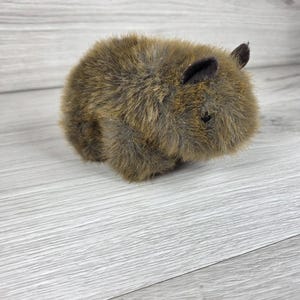 May include: A small, round plush toy shaped like a wombat, covered in shaggy, textured brown faux fur. The stuffed animal features two small, dark, pointed ears and a single visible black eye, with a compact, stout body and soft, dense fur.