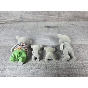 May include: Four gray toy koala figures. One koala is wearing a plaid shirt and green pants. The other three koalas are in a standing or sitting position. The toys are on a gray wooden surface.