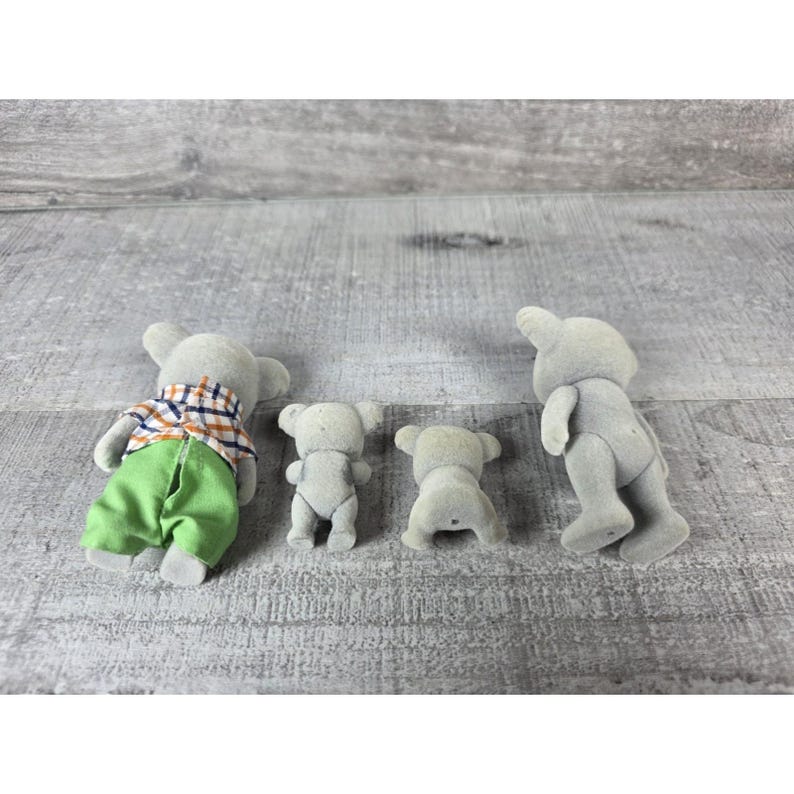 May include: Four gray toy koala figures. One koala is wearing a plaid shirt and green pants. The other three koalas are in a standing or sitting position. The toys are on a gray wooden surface.