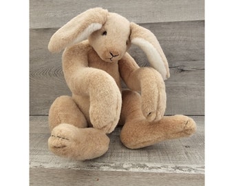May include: A beige plush rabbit toy with long ears and white fur on the inside of the ears. The rabbit is sitting with its arms crossed and its legs tucked under its body.