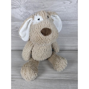 May include: A plush dog toy with shaggy tan fur, featuring long white inner ears, a dark brown nose, and a small white patch of fur above the right eye. The stuffed animal has a soft, textured appearance with floppy ears and a rounded snout.