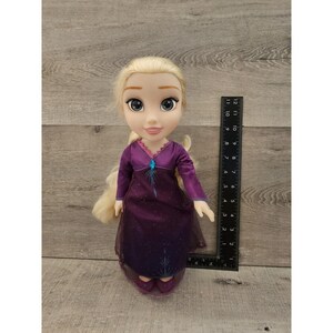 May include: A blonde doll wearing a purple dress with a blue snowflake design. The doll has blue eyes and is wearing purple shoes.