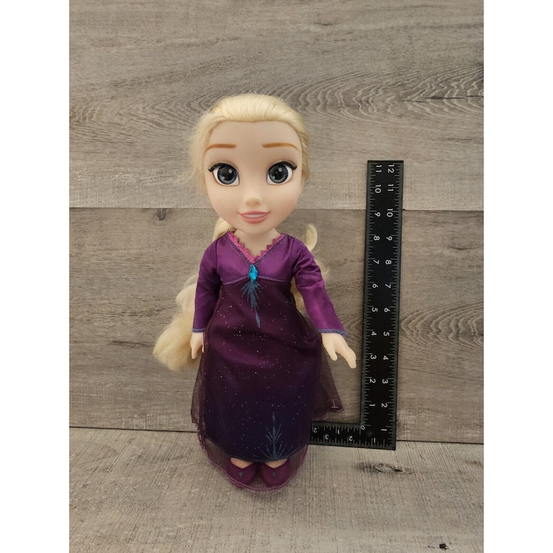 May include: A blonde doll wearing a purple dress with a blue snowflake design. The doll has blue eyes and is wearing purple shoes.