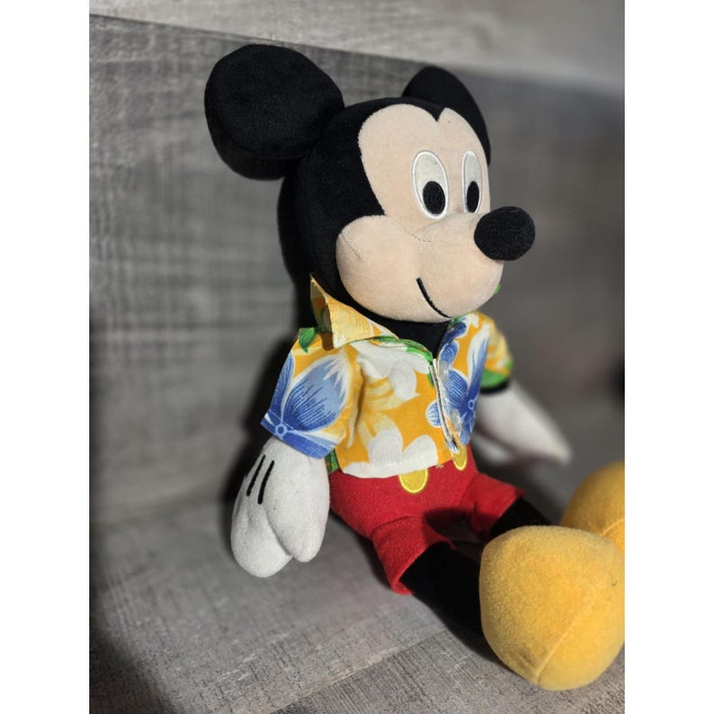 Puede incluir: Mu&ntilde;eco de peluche de Mickey Mouse con una camisa hawaiana amarilla con estampados florales azules, pantalones cortos rojos y zapatos amarillos. El mu&ntilde;eco tiene orejas negras, nariz negra y guantes blancos. El mu&ntilde;eco est&aacute; sentado.