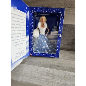 Peut inclure: Une poupée Barbie portant une robe bleue et blanche avec un châle en fourrure blanche et un motif flocon de neige. La poupée tient un manchon en fourrure blanche. La poupée est dans une boîte bleue avec un motif flocon de neige.