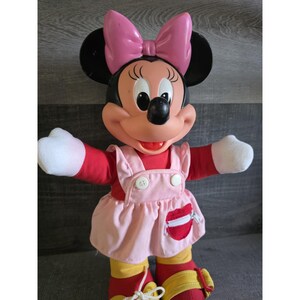 May include: A Minnie Mouse doll with a pink bow, black ears, and a red and pink outfit. The doll has white gloves, a red top, and a pink dress with white buttons. The doll is wearing red and yellow shoes.