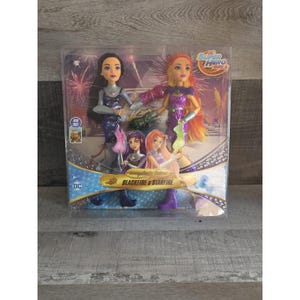May include: A packaged set of DC Super Hero Girls dolls featuring Blackfire and Starfire. The dolls are in a clear plastic case, with a colorful background. The dolls are wearing superhero outfits. The packaging includes the text "Intergalactic Sisters" and "Blackfire & Starfire".
