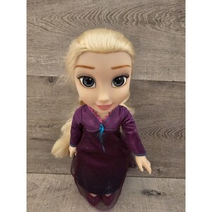 May include: A blonde doll wearing a purple dress with a blue snowflake design. The doll has a light blue gem on her chest and is wearing purple shoes.