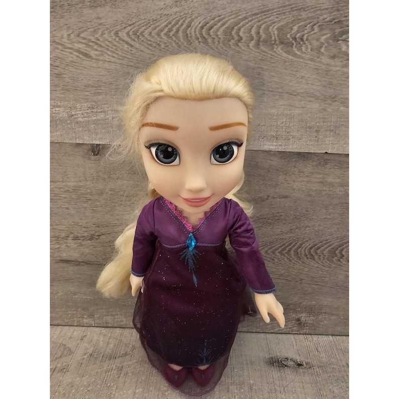 May include: A blonde doll wearing a purple dress with a blue snowflake design. The doll has a light blue gem on her chest and is wearing purple shoes.