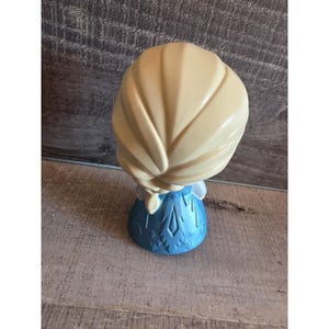 May include: A figurine of a character with blonde hair in a braid, wearing a blue dress. The figurine is viewed from the back, showcasing the details of the hair and dress design. The base of the figurine has a snowflake pattern.