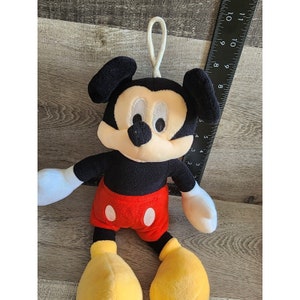 May include: A plush Mickey Mouse toy with black ears, a black body, red shorts with a white circle, and yellow shoes. The toy has a loop on the top of its head.