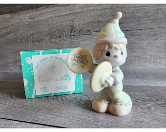 May include: A porcelain figurine of a smiling clown holding a cymbal. The figurine is wearing a green and white outfit and a green hat. The figurine is part of the Precious Moments Birthday Club.