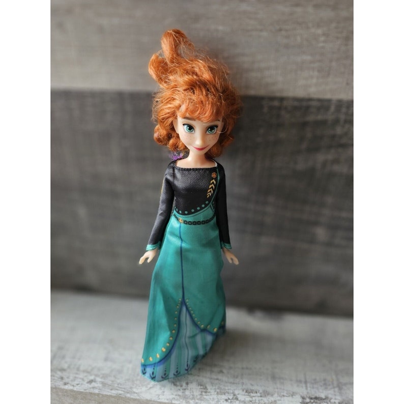 May include: A red-haired doll wearing a black and teal dress with gold accents. The doll has a friendly expression and is wearing a crown.