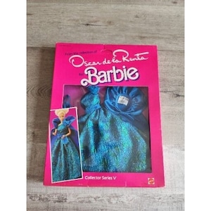 Może przedstawiać: Różowe pudełko z lalką Barbie ubraną w niebieską i zieloną sukienkę oraz niebieski szal. Na pudełku widnieje napis "Oscar de la Renta for Barbie" oraz "Collector Series V".