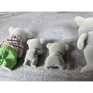 May include: Several small, gray toy koala figures are arranged on a light gray surface. One koala is wearing a plaid shirt and green pants. The other koalas are in various poses, with their backs facing the viewer.