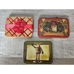 May include: Three Christmas themed tin boxes. One box has a red bow on a gold and red checkered background. Another box has a red background with a Santa Claus illustration and the text "A Merry Christmas". The third box has a green background with a cartoon moose illustration.
