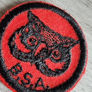 May include: A circular embroidered patch featuring a red felt background with a black stitched border. The design displays a black embroidered owl head centered on the patch, with the letters B.S.A. stitched in black at the bottom left edge.