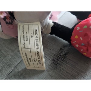 May include: A white and pink plush toy with black eyes and a pink bow. The toy has a tag that says "Surface Washable Only. Air Dry. Age:2+ Surface lavable seulement. S&eacute;cher &agrave; l'air. Age:2+ MADE IN CHINA/FABRIQU&Eacute; EN CHINE"