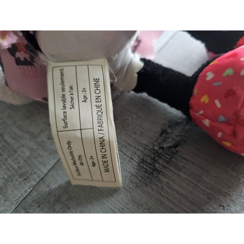 May include: A white and pink plush toy with black eyes and a pink bow. The toy has a tag that says "Surface Washable Only. Air Dry. Age:2+ Surface lavable seulement. S&eacute;cher &agrave; l'air. Age:2+ MADE IN CHINA/FABRIQU&Eacute; EN CHINE"